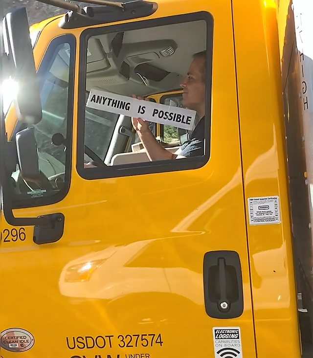 Chad Kempel holding the Anything Is Possible sign up to the window of a yellow moving truck on the drive to Eagle, Idaho, in May 2019