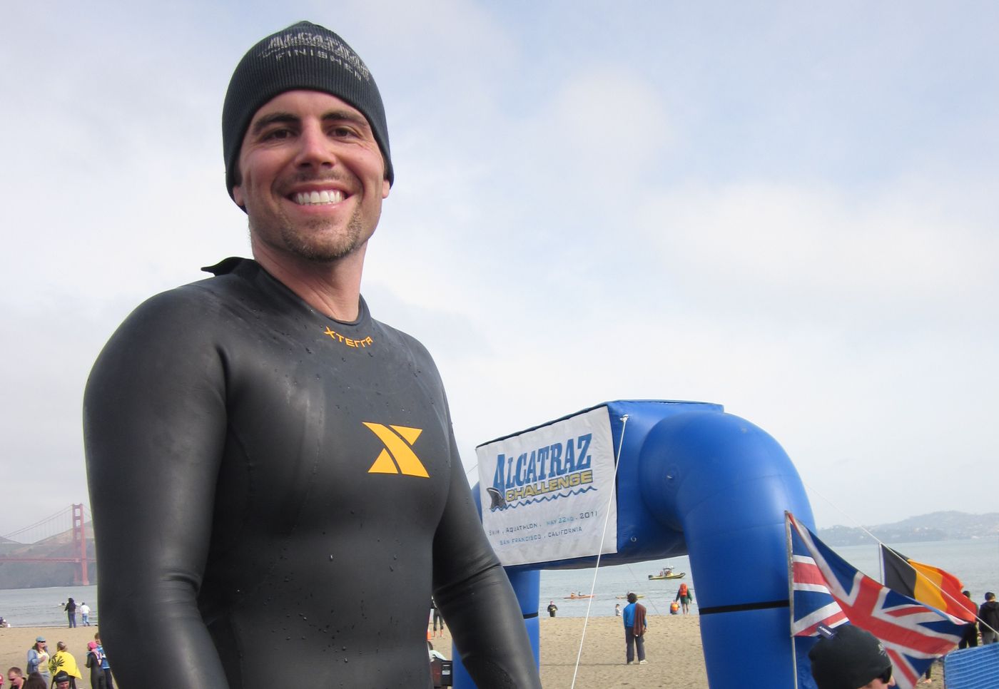 Chad Kempel smiling in his wetsuit and finisher beanie at the Alcatraz Challenge finish in San Francisco, with the Golden Gate Bridge behind him