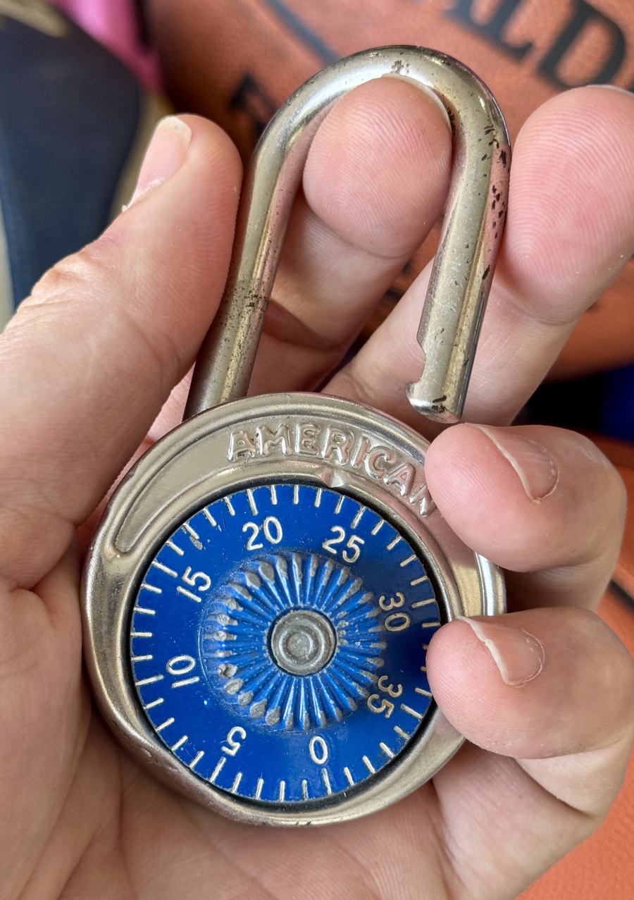 Chad Kempel's blue-dial American combination padlock from his eighth-grade gym locker, held open in his hand, the chrome shackle worn and pitted