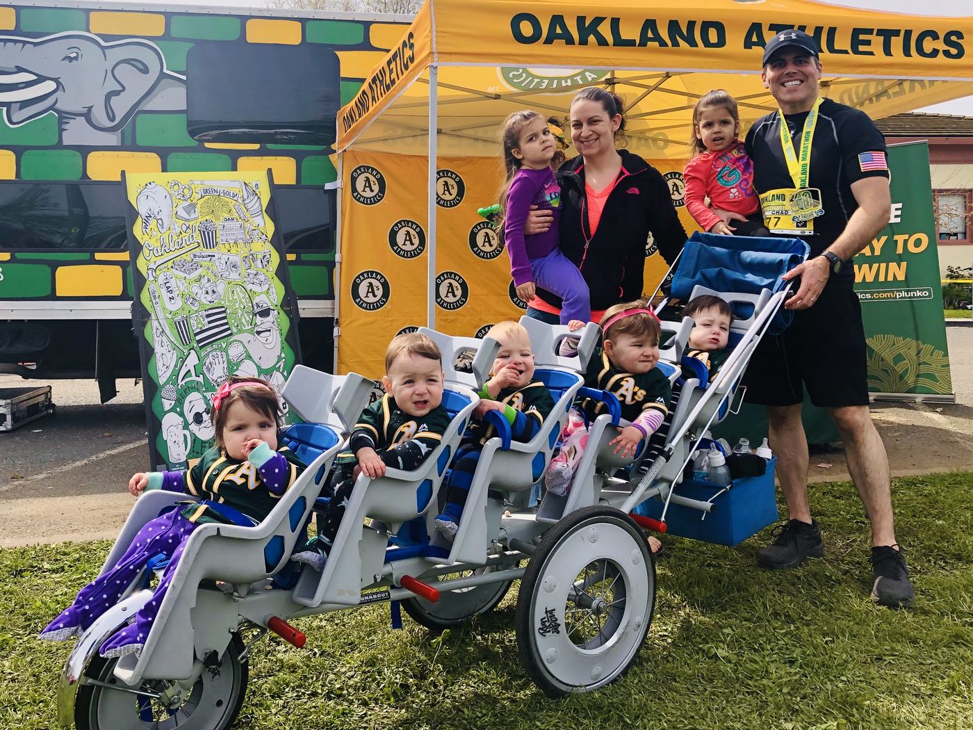 Chad Kempel in his Oakland Marathon finisher medal with his wife Amy, their two older daughters, and the five quintuplets in Oakland A's jerseys in the stroller