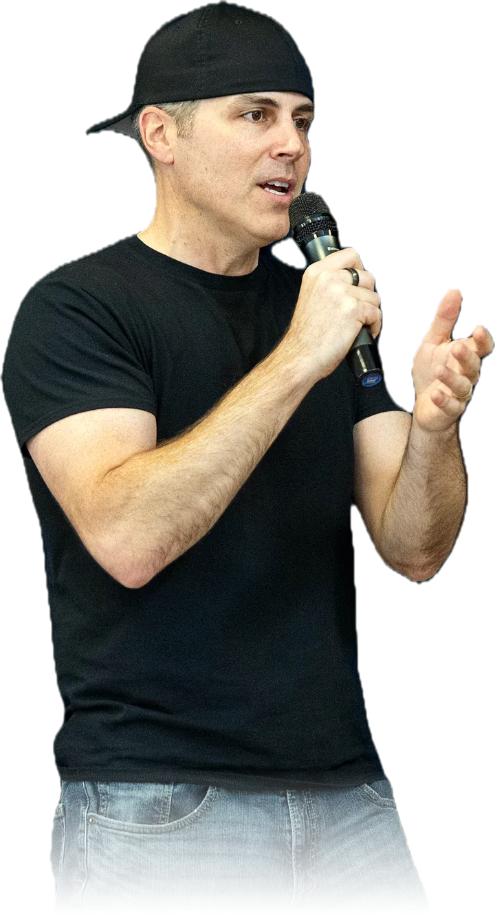 Chad Kempel, high school assembly speaker, talking into a handheld microphone in a black T-shirt and backwards cap