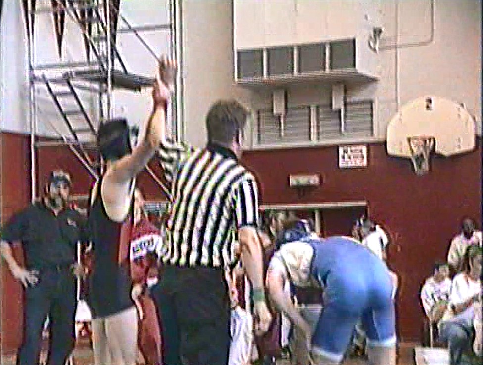 Chad Kempel in tenth grade, a referee raising his arm after a wrestling match in the Arroyo High School gym, with red doors and a basketball hoop on the wall behind them