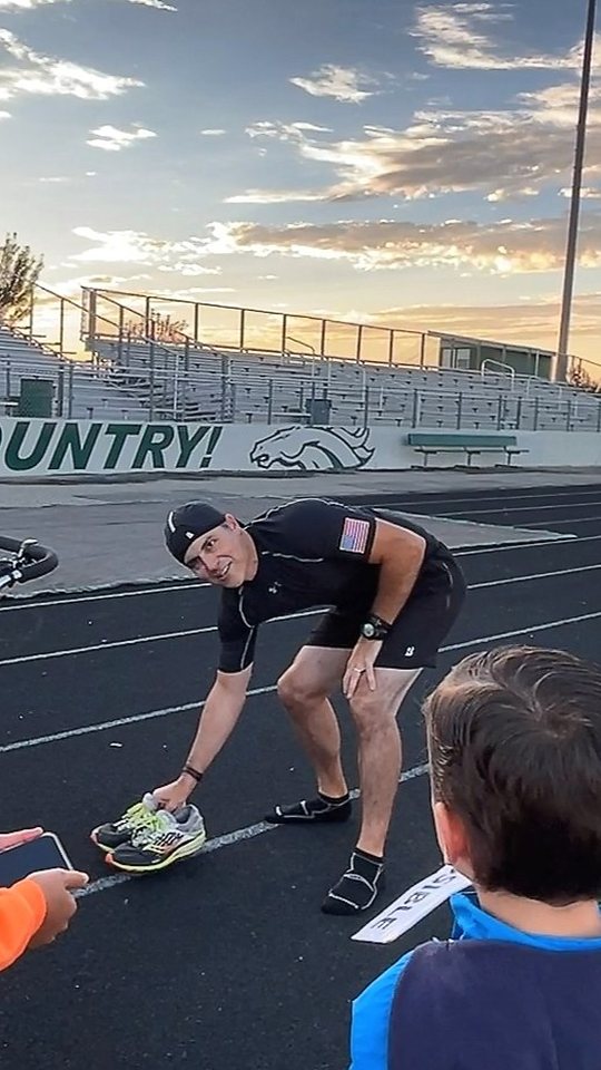 Chad Kempel in socks bending down to set his running shoes on the Eagle High School track after his final stroller world record, with the Mustang Country wall behind him