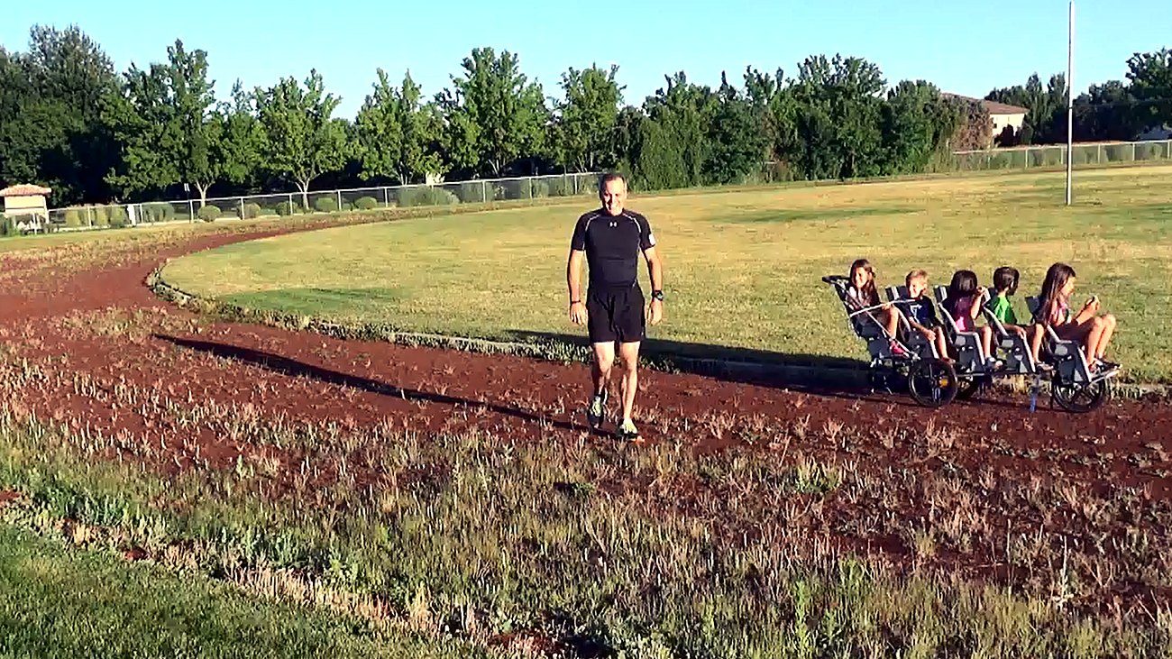 Chad Kempel walking back along the red cinder track at Eagle Middle School in Eagle, Idaho, with weeds growing through the loose surface and the five-seat stroller parked behind him after a failed 1K record attempt