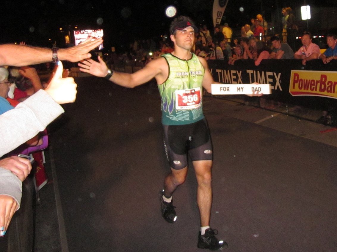 Chad Kempel high-fiving spectators at the Ironman Canada finish while holding the other side of the sign, which reads For My Dad