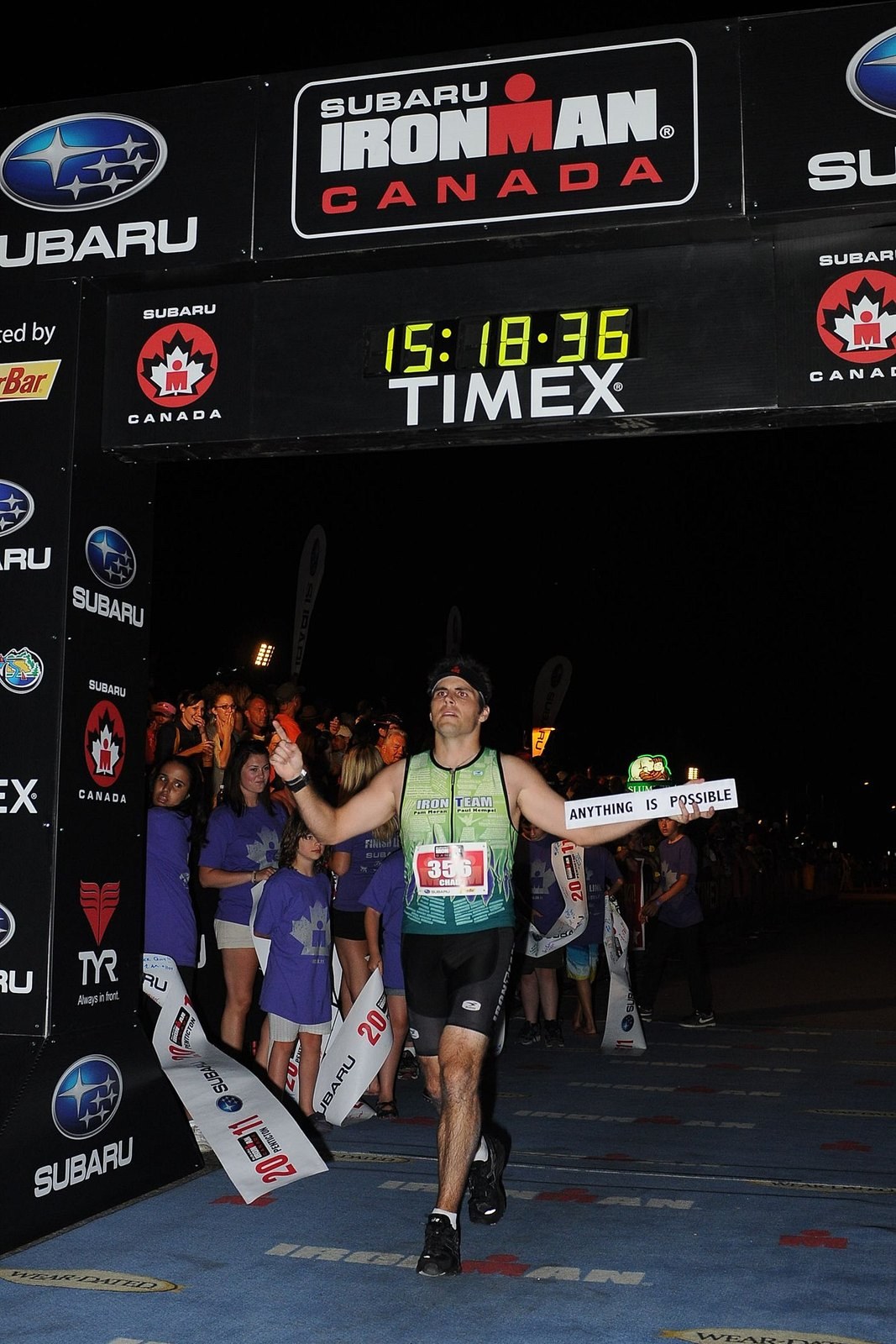 Chad Kempel crossing the Ironman Canada finish line at night holding a sign that reads Anything Is Possible, with the clock showing 15:18:36