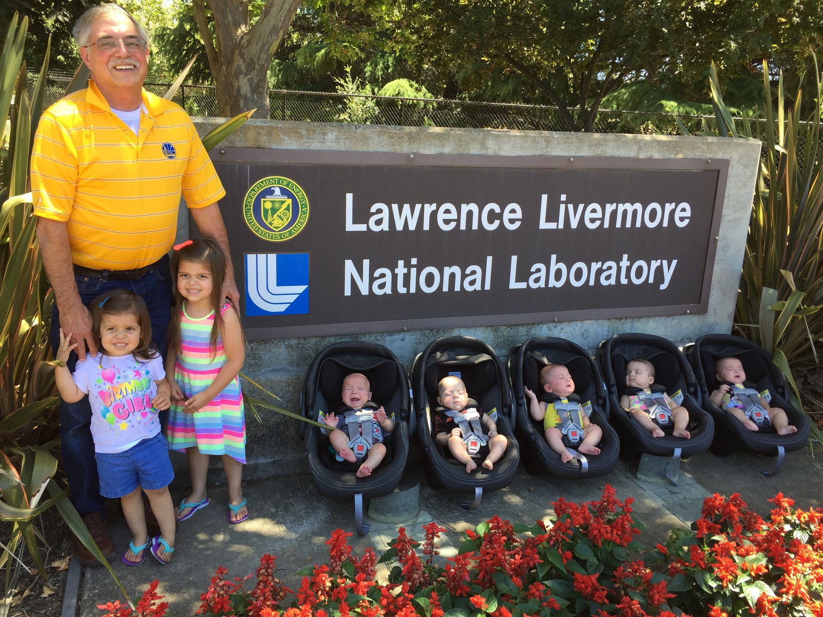 Paul Kempel with his seven grandchildren, two girls standing beside him and five infants in car seats, at the Lawrence Livermore National Laboratory sign