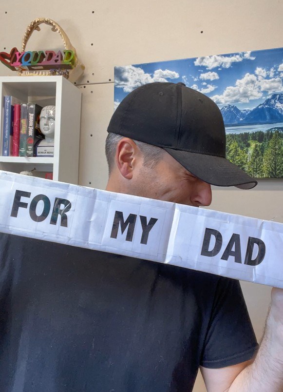 Chad Kempel in a black cap holding the cardboard sign across his face so it reads For My Dad
