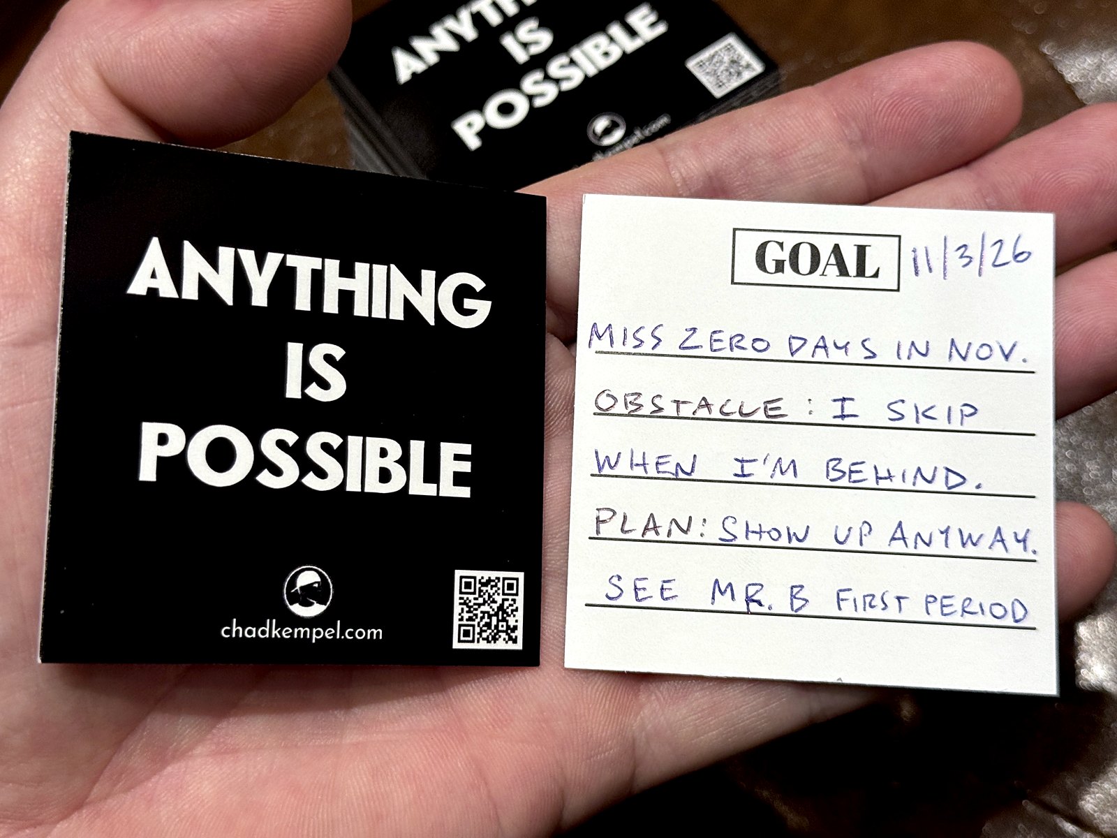 Two goal cards in a hand. The black front says Anything Is Possible with a QR code. The white back says GOAL and, in Chad's handwriting, a sample: miss zero days in November; obstacle, I skip when I'm behind; plan, show up anyway, see Mr. B first period.