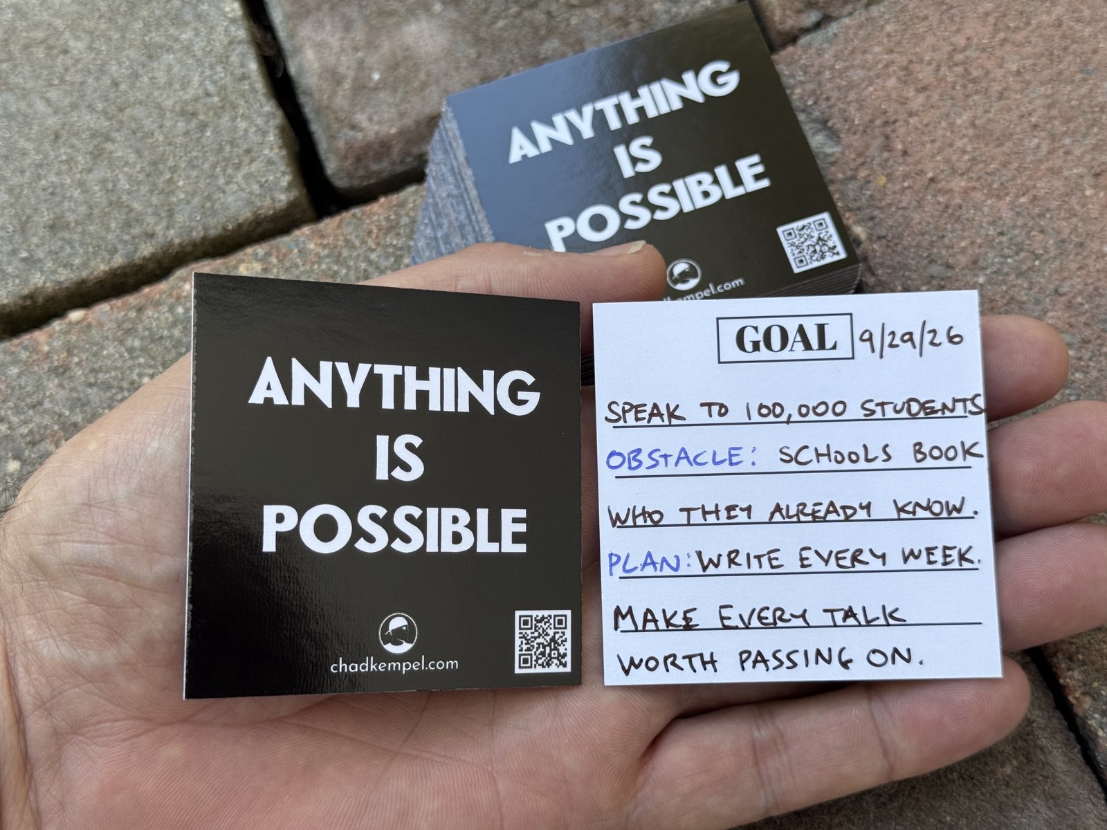 A hand holding two goal cards on a brick walkway: a black card that reads Anything Is Possible with chadkempel.com and a QR code, and a white card with GOAL at the top and a handwritten goal, obstacle and plan