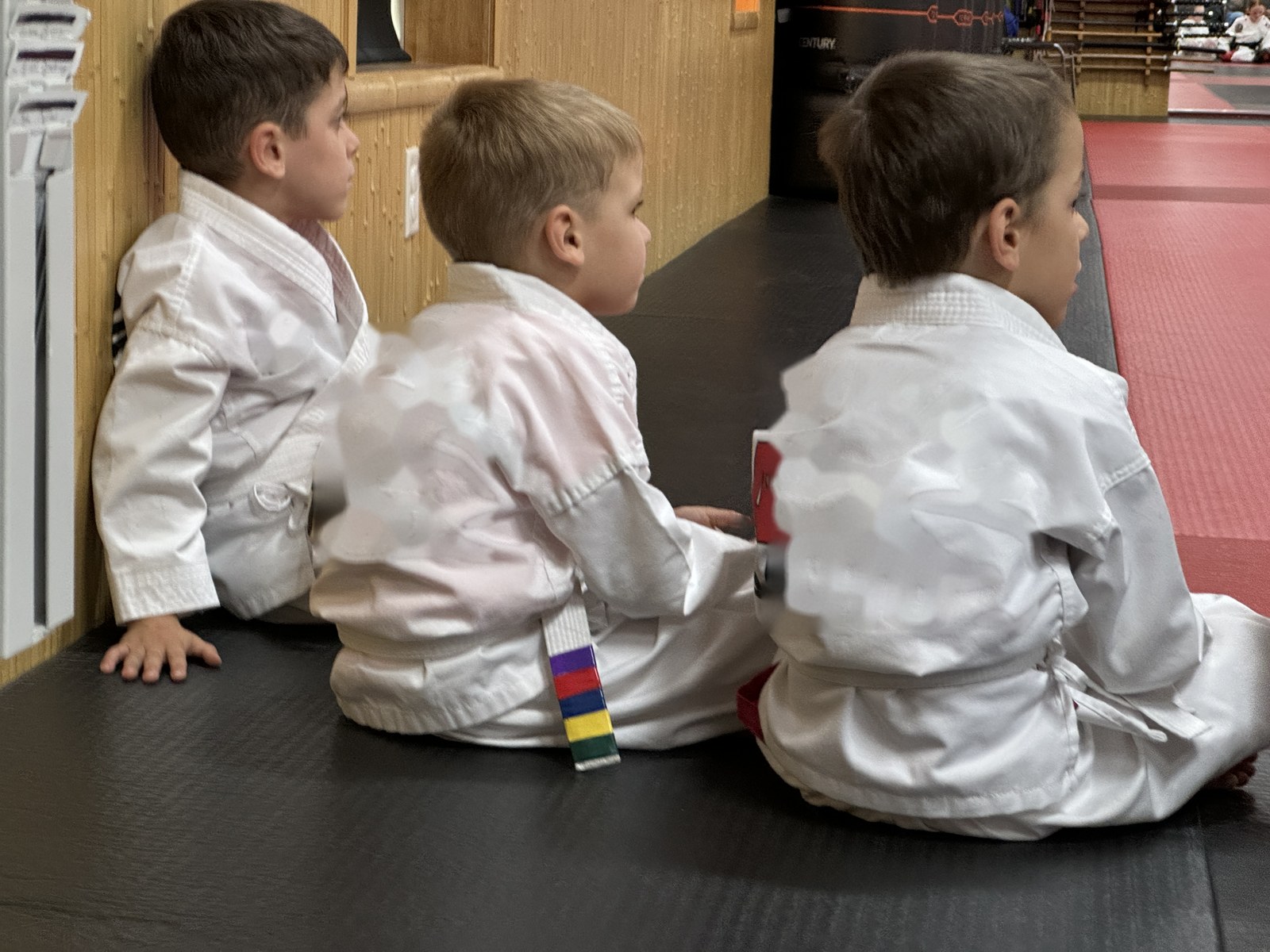 Three young boys in white martial arts uniforms sitting side by side on a mat, watching class