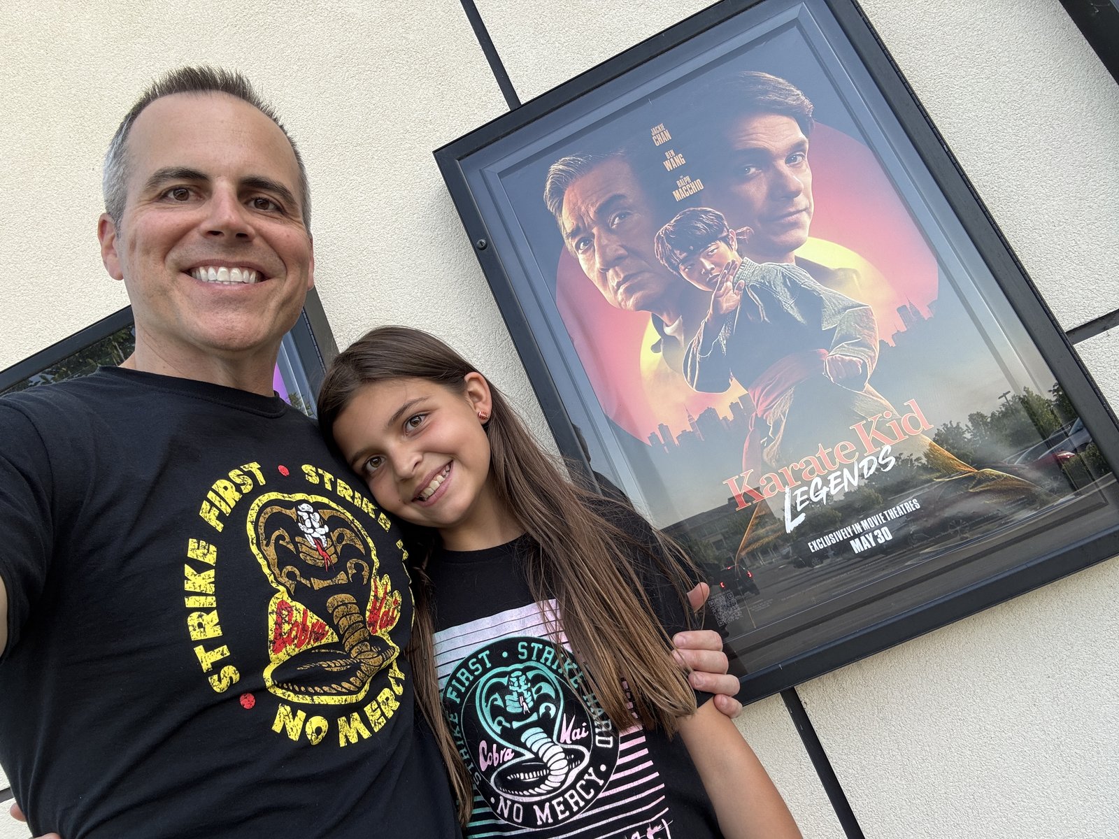 Chad Kempel and his daughter, both in Cobra Kai shirts, smiling in front of a Karate Kid: Legends movie poster