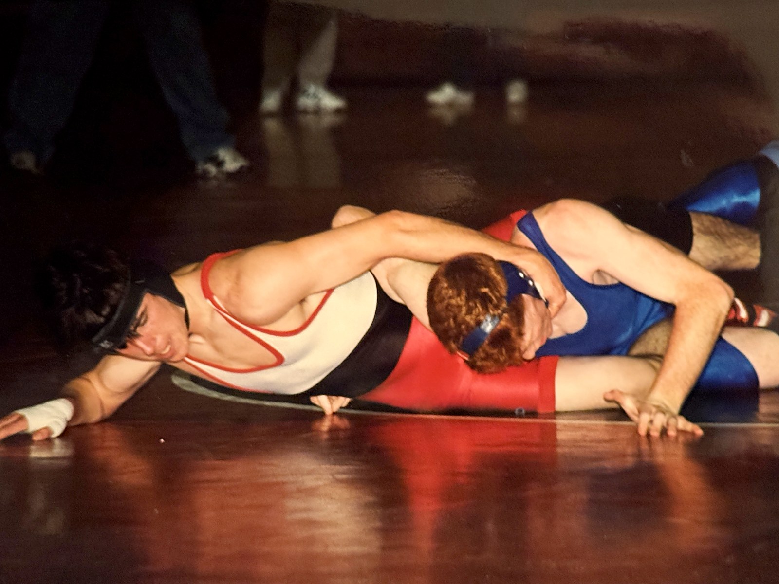 Chad Kempel in a white, red and black wrestling singlet and black headgear controls an opponent in a blue singlet on a dark gym mat