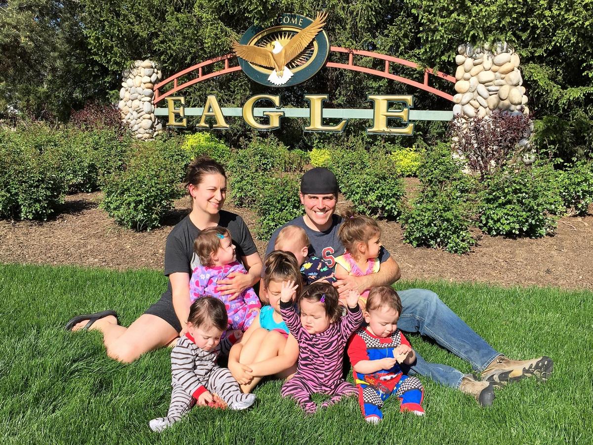 Chad and Amy Kempel sitting on the grass with their seven children, the quintuplets a year old, in front of the Welcome to Eagle sign in Eagle, Idaho, on the day they moved in, May 12, 2019
