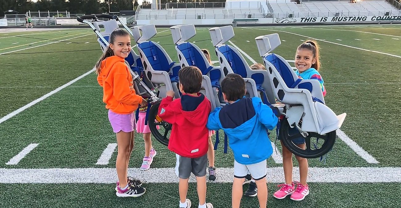 Five of Chad Kempel's children lifting the empty five-seat stroller together on the Eagle High School football field, with the This Is Mustang Country sign on the bleachers behind them and two adults walking laps on the track at the top left