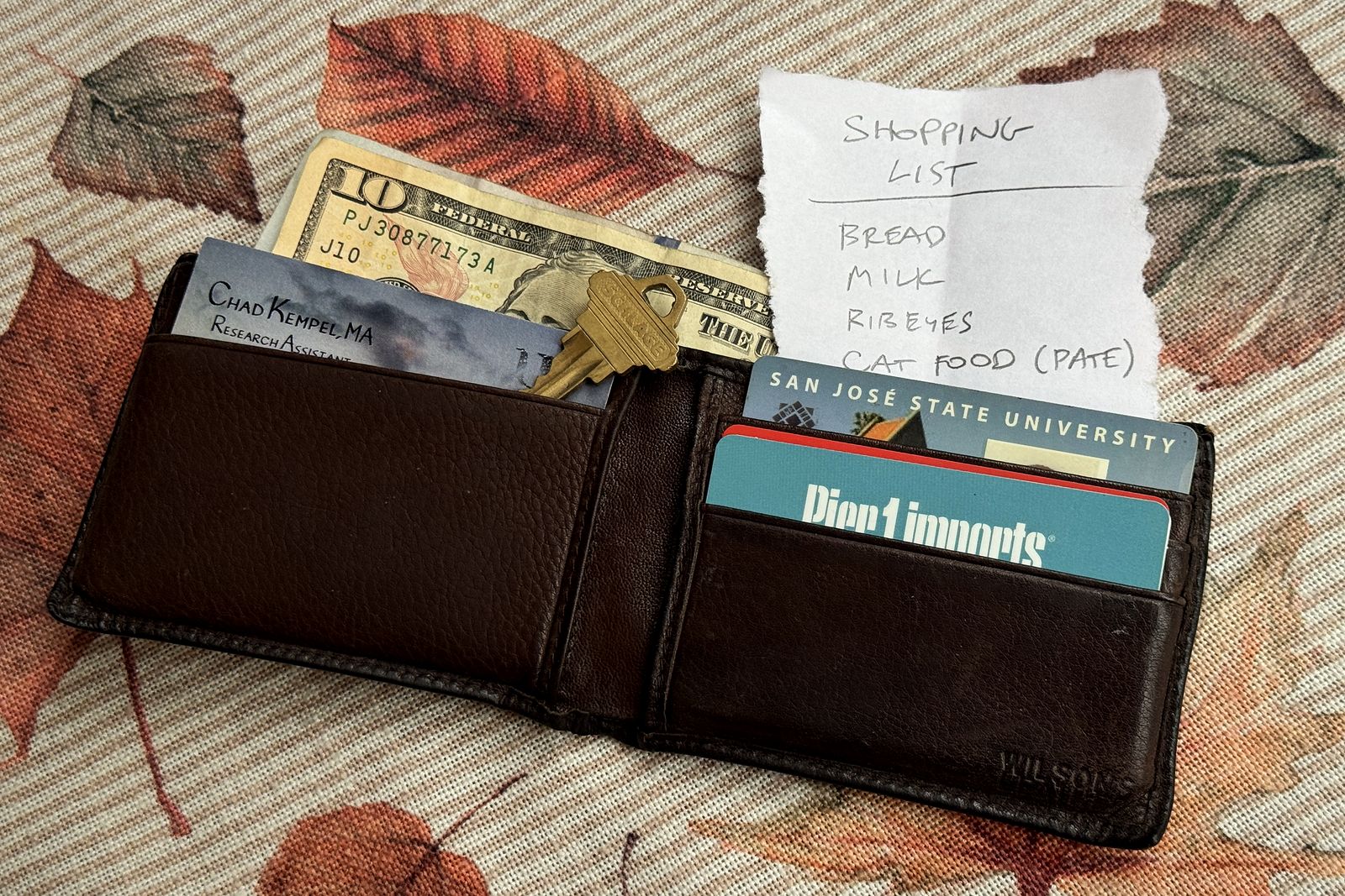 An open brown leather wallet holding a ten-dollar bill, a house key, business cards and a handwritten shopping list