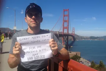 Chad Kempel on the Golden Gate Bridge at the end of his 100-mile run, holding a sign with his start and finish times, dedicated to his wife