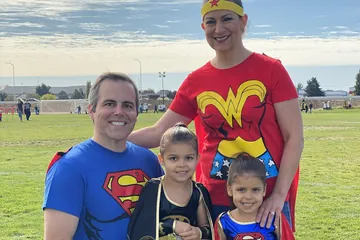 Chad Kempel and his wife in superhero shirts with Savannah and Avery in costume at the Superhero Obstacle Fun Run