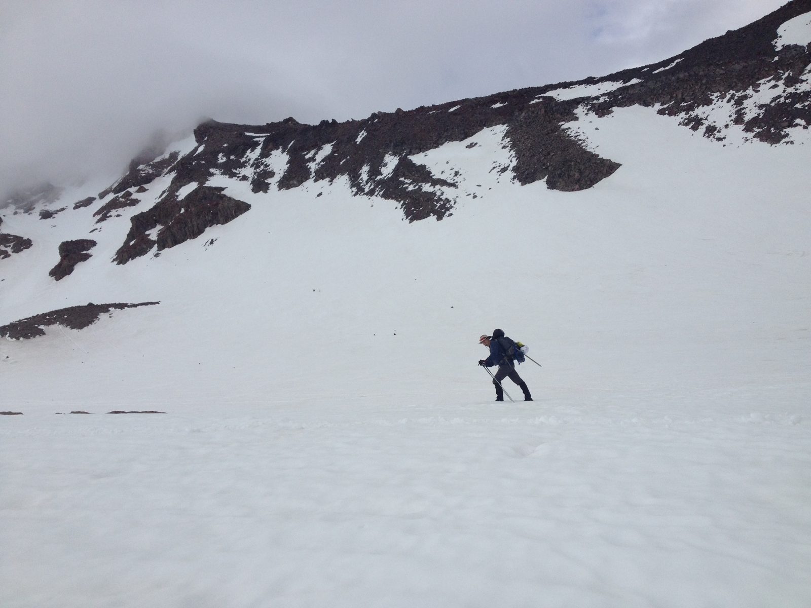 Jeff climbing a wide snowfield on Mt. Shasta with trekking poles and a heavy pack, rock ridges and clouds above