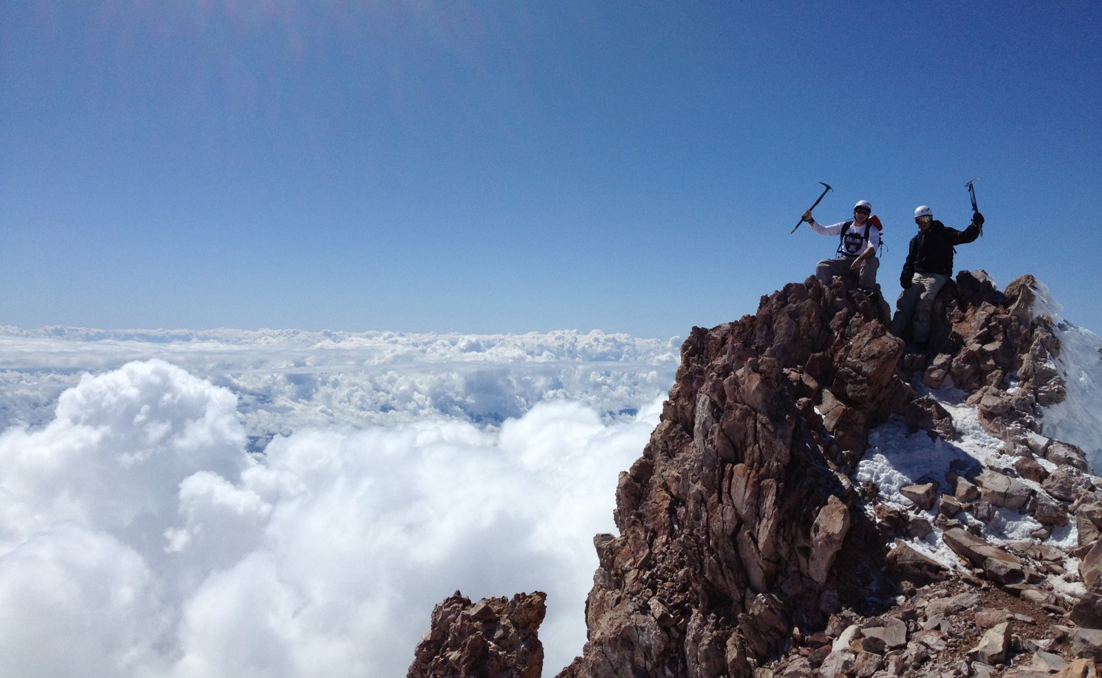 Chad Kempel, left, and his friend Jeff raising ice axes on the rocky summit of Mt. Shasta with clouds below them