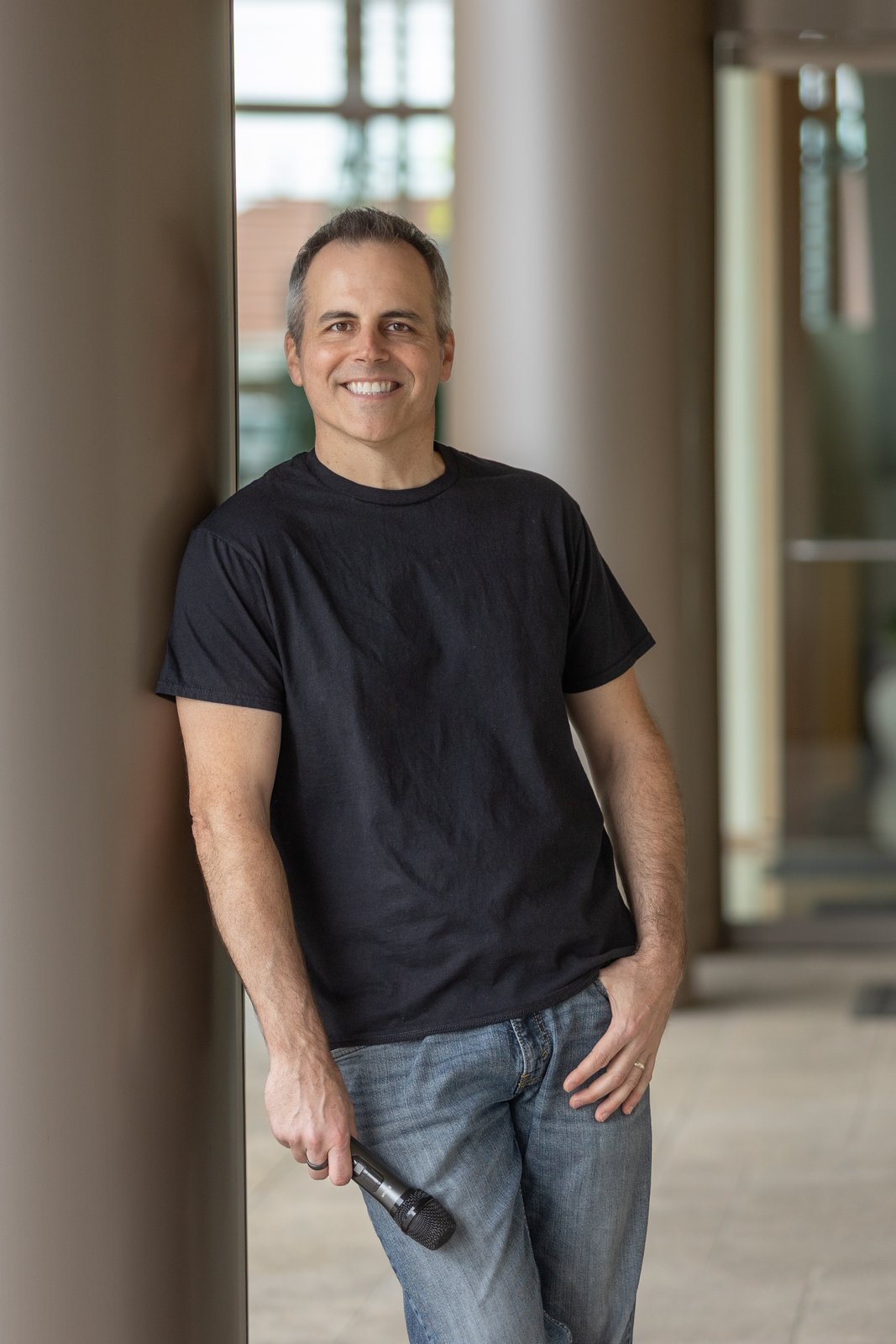 Chad Kempel, smiling portrait in black T-shirt holding a microphone
